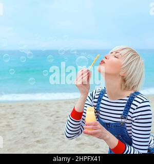 portrait de belle jeune jolie, heureuse, mignonne, blonde femme dans une chemise rayée, salopette denim avec bulles de savon près de la mer Banque D'Images