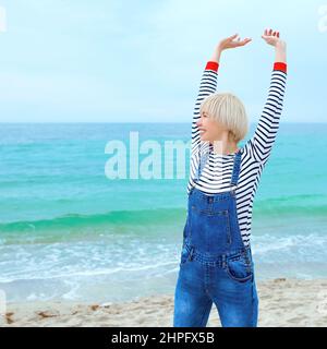 belle jeune femme blanche blonde en vacances en blouse rayée, baskets et denim en général sur la plage à côté de l'incroyable fond bleu de mer. Banque D'Images