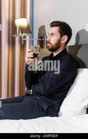 homme barbu tenant un verre de brandy et assis dans le lit de l'hôtel Banque D'Images