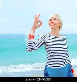 belle jeune femme blonde caucasienne en vacances avec chemisier rayé, baskets et denim dans l'ensemble sur la plage par le fond bleu de la mer. Heureux d'être Banque D'Images