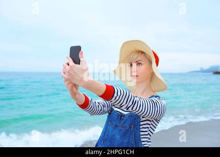 belle jeune femme blonde caucasienne en vacances à chapeau de paille, chemisier rayé nad denim salopettes faisant selfie sur smartphone près de l'incroyable mer bleue Banque D'Images
