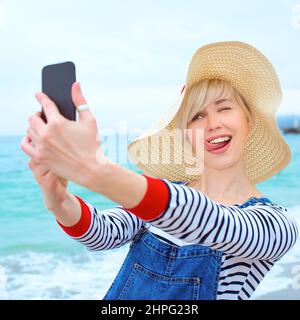 belle jeune femme blonde caucasienne en vacances à chapeau de paille, chemisier rayé nad denim salopettes faisant selfie sur smartphone près de l'incroyable mer bleue Banque D'Images