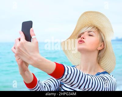 belle jeune femme blonde caucasienne en vacances à chapeau de paille, chemisier rayé nad denim salopettes faisant selfie sur smartphone près de l'incroyable mer bleue Banque D'Images