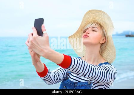 belle jeune femme blonde caucasienne en vacances à chapeau de paille, chemisier rayé nad denim salopettes faisant selfie sur smartphone près de l'incroyable mer bleue Banque D'Images