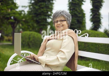 Bonne femme âgée avec un ordinateur portable assis sur le banc de parc regardant la caméra Banque D'Images