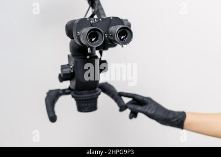 Gros plan de la main du dentiste dans un gant noir touchant le microscope dentaire - équipement innovant pour le traitement des dents. Concept de soins de santé bucco-dentaire, clinique de stomatologie professionnelle et moderne. Banque D'Images