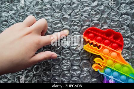 Jouet pop il dinosaure couleurs arc-en-ciel sur un arrière-plan de bulle avec des lettres multicolores - Pop il. Le doigt d'enfant pousse la bulle, dino la regarde. Copier sp Banque D'Images