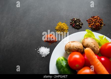 Une assiette blanche de légumes frais : champignons, tomates, carottes et herbes, autour de laquelle diverses épices sont dispersées en piles. Sur fond noir Banque D'Images