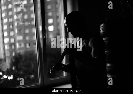 Silhouette de boxeur musclé méconnaissable enveloppant le ruban de boxe de défense autour des bras avant de se battre dans le centre de fitness avec intérieur sombre. Banque D'Images