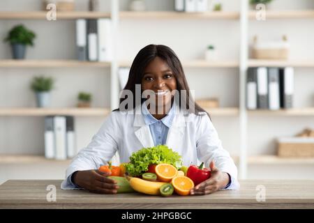 Portrait d'un diététicien afro-américain heureux en blouse de laboratoire, assis avec des fruits et des légumes frais sur la table Banque D'Images