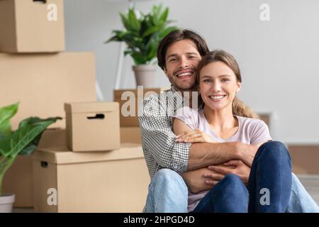 Jeune couple caucasien excité satisfait dans des hugs décontractés, assis parmi des boîtes en carton avec des effets personnels dans la chambre Banque D'Images
