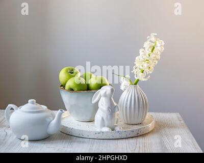 Une jacinthe blanche dans un vase à 70s cannelures à côté d'un plateau avec un grand bol de pommes vertes, un lapin en porcelaine de Pâques et une théière se trouvent sur la table. Min Banque D'Images