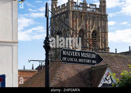 Un panneau noir avec des lettres blanches pointant vers le Musée d'histoire locale de Malvern avec la tour carrée du Grand prieuré de Malvern derrière. ROYAUME-UNI Banque D'Images