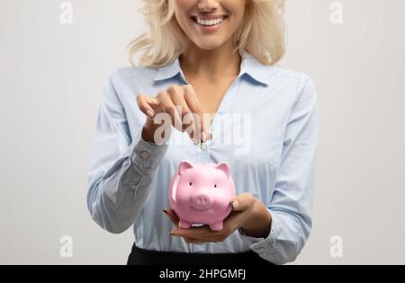 Concept d'épargne et d'investissement. Femme d'affaires mettant l'argent dans la banque de porc rose, fond gris clair de studio, récolte Banque D'Images