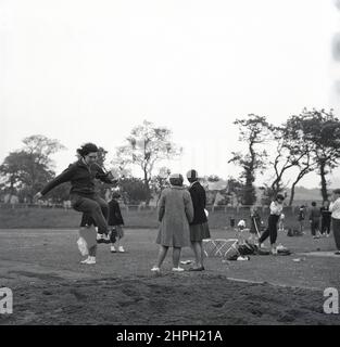 1960s, historique, une jeune athlète féminine dans sa combinaison d'athlétisme en coton faisant un saut d'entraînement ou d'échauffement dans le long saut, un événement d'athlétisme à une compétition sportive, Fife, Scotand, Royaume-Uni. La vitesse, la force et l'agilité sont nécessaires dans le long saut, où l'objectif est de sauter le plus loin possible d'un point de décollage lors de la course. Un événement masculin dans les premiers Jeux Olympiques modernes de 1896, les femmes ont pris part pour la première fois au long saut olympique à Londres en 1948. Banque D'Images