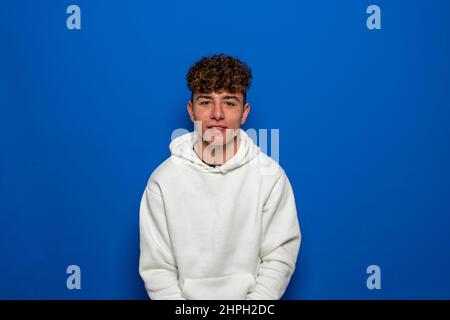 Sérieux beau jeune homme caucasien avec des cheveux bouclés regardant la caméra sur un fond bleu dans un sweat-shirt blanc. Banque D'Images