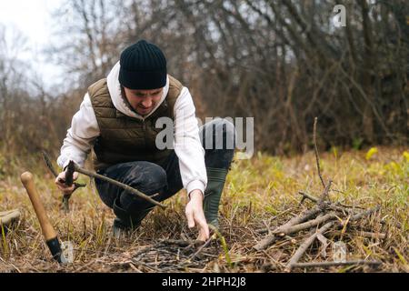 Vue de face du voyageur perdu homme préparant du bois de chauffage au feu de camp pour garder au chaud et cuisiner à l'extérieur sur couvert froid jour. Banque D'Images