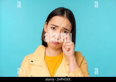 Gros plan de triste fille asiatique pleurant, essuyant des larmes et des tergiverser, regardant bouleversé et sombre, posant sur fond bleu Banque D'Images