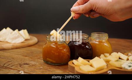 Pots de confiture et de miel faits maison dans des pots en verre et variété de fromages sur une table en bois. Main tenant une brochette avec du fromage. Assiette de fromages pour les hors-d'œuvre Banque D'Images