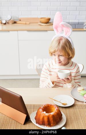 Portrait vertical d'un garçon mignon portant des oreilles de lapin tout en prenant le petit déjeuner le matin de Pâques et en utilisant une tablette dans la cuisine Banque D'Images