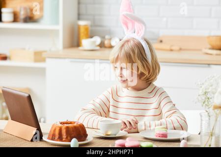 Vue latérale portrait d'un garçon mignon portant des oreilles de lapin tout en prenant le petit déjeuner le matin de Pâques et en utilisant une tablette dans la cuisine Banque D'Images