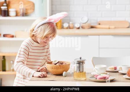 Vue latérale portrait d'un joli garçon blond portant des oreilles de lapin à Pâques et mangeant des œufs de chocolat doux, espace copie Banque D'Images