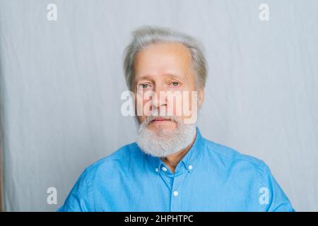 Portrait en gros plan d'un homme âgé sérieux aux cheveux gris qui regarde l'appareil photo sans émotion sur un arrière-plan blanc isolé. Banque D'Images