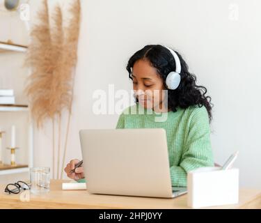 Une femme afro-américaine prend des notes tout en écoutant un podcast audio dans un casque. Cours en ligne Banque D'Images