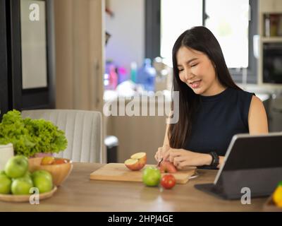 Femme asiatique millénaire hacher des pommes vertes et rouges, préparer un brunch sain tout en regardant la recette sur un comprimé dans la cuisine. Banque D'Images