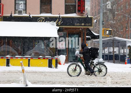 Un messager de livraison de nourriture à vélo sur un e-vélo sur une rue enneigée et lushy à New York avec un restaurant libre et hors d'affaires à louer à l'arrière-plan Banque D'Images