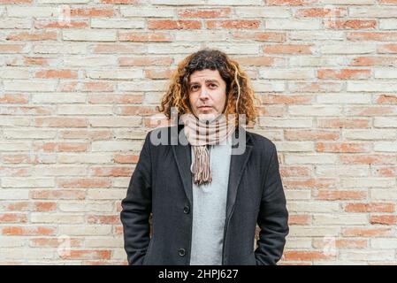 Homme d'âge moyen en manteau élégant et décontracté avec écharpe et cheveux bouclés tenant les mains dans les poches. Il regarde la caméra tout en se tenant contre le mur de briques sur la rue de la ville Banque D'Images