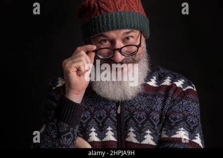 L'homme caucasien à barbe mûre semble slicieusement dans l'appareil photo avec des lunettes sur son visage, l'homme en chapeau de père noël rouge, fond noir Banque D'Images