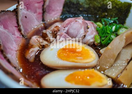un gros plan d'un bol de soupe avec de la saumure marinée, un œuf demi-cuit, des tranches de porc et de viande, du nori, de la graisse et de la bonté d'oignon vert frais et haché. Banque D'Images