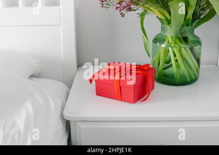 Bouquet de tulipes et boîte cadeau rouge sur la table de nuit près du lit dans la chambre. Concept du jour des femmes, jour des mères Banque D'Images