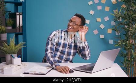 Homme afro-américain à l'écoute de potins et de rumeurs à l'ordinateur portable, à la recherche d'informations confidentielles et à l'écoute d'écoutes. Jeune personne qui entend une conversation privée secrète. Banque D'Images