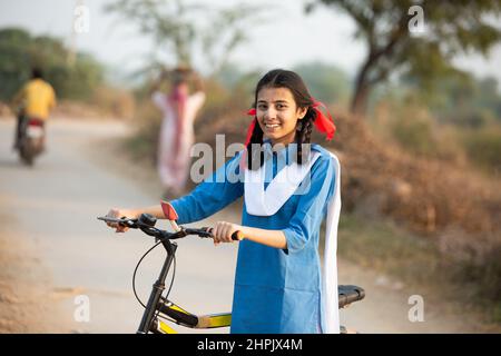 Portrait d'une jeune fille indienne rurale heureuse portant un uniforme scolaire bleu debout avec un vélo dans la rue du village. Banque D'Images