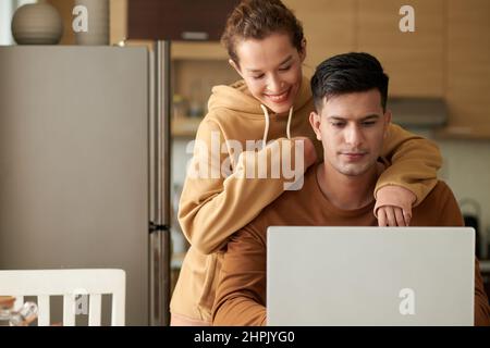 Jeune homme travaillant sur un ordinateur portable assis à la table dans la cuisine avec une femme heureuse l'embrassant de derrière Banque D'Images