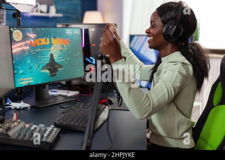 Bonne femme afro-américaine jouant avec un casque gagnant jeux vidéo compétition parlant dans le microphone avec les joueurs. Un joueur enthousiaste jouant à des jeux de tir spatial en utilisant un ordinateur RVB. Diffusion en direct Banque D'Images