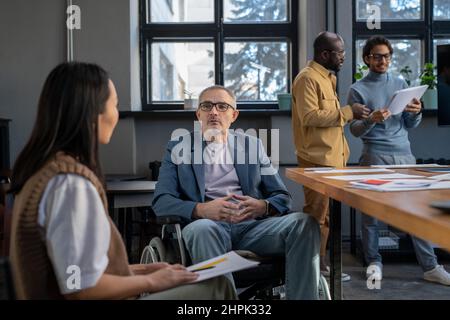 Homme d'affaires mature en fauteuil roulant à l'écoute de la jeune collègue féminine lors d'une réunion de travail, assis contre deux collègues interculturels Banque D'Images