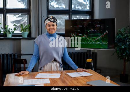 Jeune coach d'affaires contemporain debout à table pendant la présentation des données financières et regardant la caméra dans la salle de réunion Banque D'Images