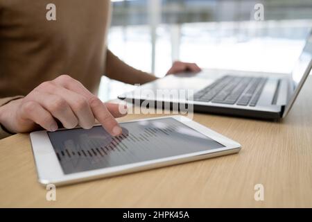 Main de jeune homme d'affaires pointant sur les données sur l'écran d'une tablette tout en analysant les graphiques pendant le travail sur les nouveaux projets d'affaires par table Banque D'Images