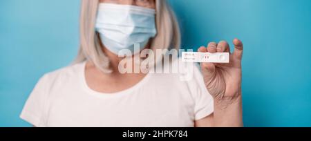 Femme dans un masque médical de protection tenant un test d'antigène avec un résultat positif pour la pandémie de COVID-19 posant sur fond bleu clair Banque D'Images