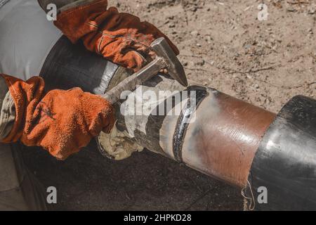 Soudeur avec un marteau en main et des mitaines de travail enlève les scories après le travail de l'industrie de soudage le tuyau de chauffage principal d'alimentation en eau de polyéthylène sur la construction Banque D'Images