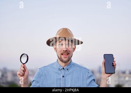 Mignon barbu homme heureux expression regardant l'appareil photo. Homme portant un Jean chemise et chapeau tenant la loupe et le smartphone dans les mains à la vue de la ville comme détective, enquêteur ou concept de recherche Banque D'Images