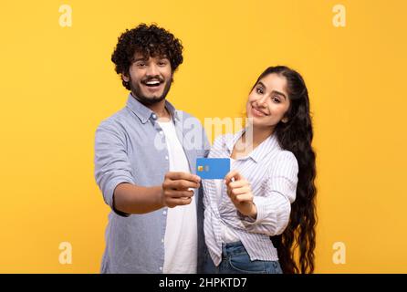 Couple indien heureux avec carte de crédit faisant la promotion des services bancaires, encourageant les achats sans contact sur fond de studio jaune. Happ homme et femme excités Banque D'Images