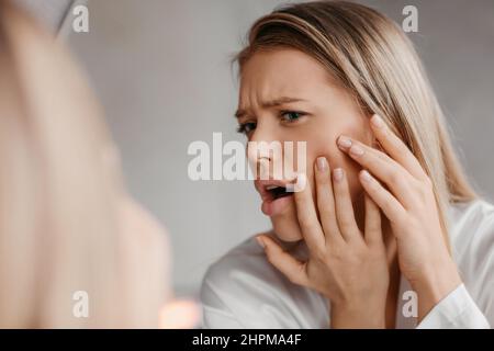 Une dame malheureuse avec le bouton-pression sur la joue tout en se tenant près du miroir dans la salle de bains à la maison, jeune femme souffrant d'acné Banque D'Images