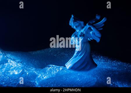 Photo de l'ange de glace miniature bleu jouant le luth et debout sur une surface de glace fissurée sur fond sombre. Banque D'Images