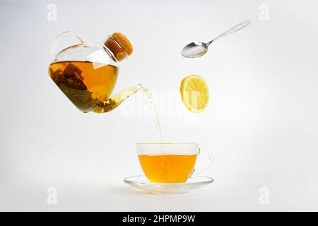 Le thé s'écoule d'une bouilloire transparente dans une tasse transparente. Une tranche de citron et une cuillère tombent Banque D'Images