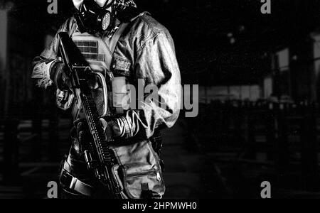 Photo en noir et blanc d'un soldat urbain en tenue militaire tactique et masque à gaz debout avec un fusil et un masque à gaz sur fond sombre d'usine. Banque D'Images