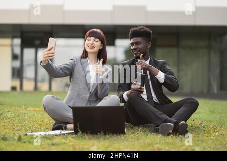 Les jeunes collègues d'affaires s'amusent à l'extérieur, s'assoyent à l'extérieur du bureau et prennent un selfie. Joyeuse femme caucasienne et son collègue africain masculin assis sur l'herbe devant le bureau et de faire des photos Banque D'Images
